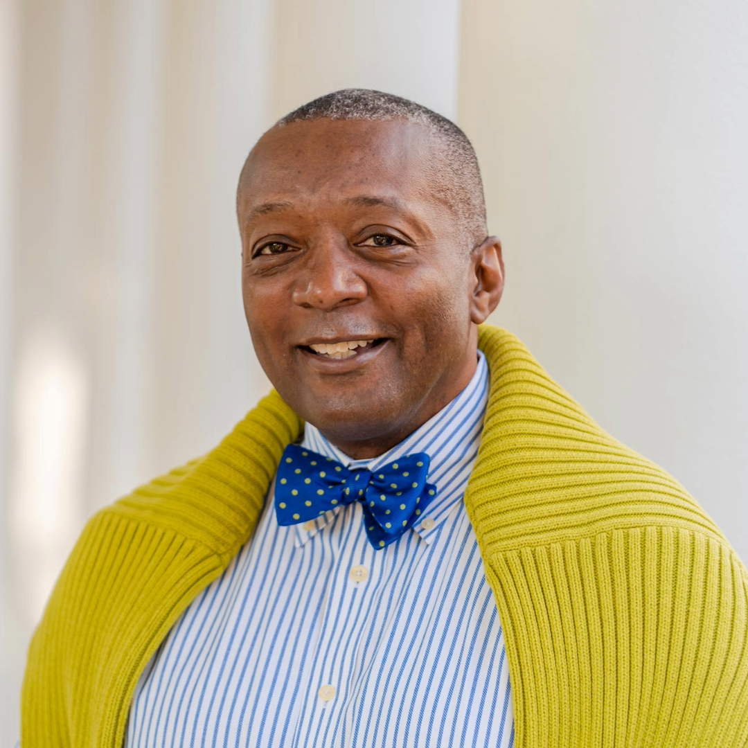 Image of Cedric Rucker wearing a blue striped button down with a navy blue bow tie. Through over his shoulders is a neon green/yellow sweater.